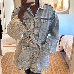 Oversized jeansjacka med bälte - Snygg oversized jeansjacka i ljusblå denim med knappar framtill, stora fickor och bälte i midjan. Jackan har klassisk krage, långa ärmar och en loose fit som ger en avslappnad vibe. Perfekt för lager-på-lager och streetstyle.