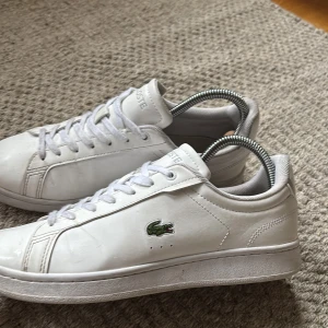 Vita sneakers från Lacoste storlek 39 - Snygga vita sneakers från Lacoste med klassisk låg profil och rund tå. Skorna har snörning, platt sula och är tillverkade i skinn. På sidan finns den ikoniska gröna krokodilen broderad och Lacoste-logga på plösen och hälen. Perfekt för dig som gillar stilrena och cleana sneakers. Kvalité 9,8/10 använd ett få tal gånger. Nytt skick ❗️❗️