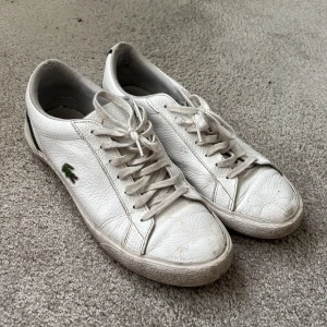 Vita sneakers från Lacoste i skinn - vita sneakers från Lacoste med klassisk krokodillogo på sidan. Skorna har snörning, rund tå och platt sula i gummi. Ovandelen är i vitt skinn med svarta detaljer på hälen. Perfekt för dig som gillar stilrena och sportiga sneakers. Stl 44