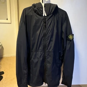 Svart vindjacka från Stone Island - Snygg svart vindjacka från Stone Island med ikonisk patch på ärmen. Jackan har huva med vit kant och i storlek M men passar L också, dragkedja framtill och två fickor med dragkedja. Tillverkad i lätt och slitstark nylon, perfekt för blåsiga dagar. Enkel och stilren design som passar till det mesta. Kan tänka mig att byta mot något annat Stone Island. OBS SKICKAS EJ! Bara avhämtning i Göteborgsområdet och säljs utan patch! Mvh