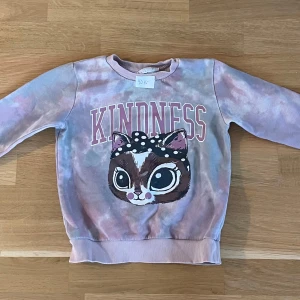 Pastellfärgad sweatshirt KappAhl 110/116 - Superfin sweatshirt från KappAhl i storlek 110/116. Tröjan har pastellfärgat batikmönster i lila, rosa och beige toner, trycket 'KINDNESS' och en söt katt med prickig rosett framtill. Mjuk mudd vid ärmslut och hals. Perfekt för dig som gillar gulliga prints!