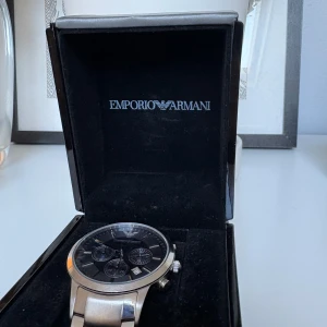 Emporio Armani silverfärgad klocka - Stilren klocka från Emporio Armani med silverfärgat armband i metall och svart urtavla. Klockan har tre mindre urtavlor, datumvisning och tydliga indexmarkeringar. Snygg och modern design som passar till många olika outfits.