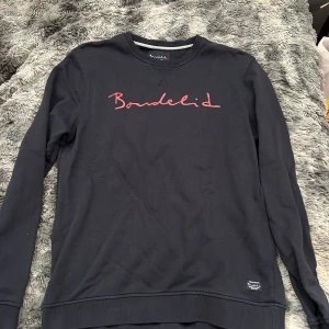 Svart sweatshirt från Bondelid M - Svart sweatshirt från Bondelid med röd logga broderad över bröstet. Klassisk rund halsringning och långa ärmar. Tröjan är i mjuk bomull och har ribbade muddar vid ärmslut och nederkant. Perfekt för en chill och stilren look.