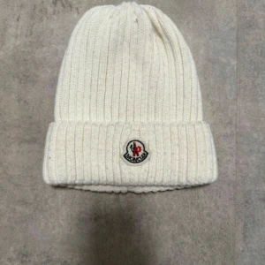 Vit ribbstickad mössa från Moncler - Säljer en snygg vit ribbstickad mössa från Moncler med uppvikt kant och klassisk logga framtill. Mössan är gjord i mjukt och varmt material, perfekt för kyliga dagar. Enkel och stilren design som passar till det mesta.