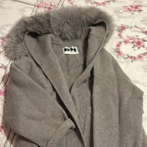 Grå kappa med fuskpäls från Dennis Maglic - Snygg grå kappa från Dennis Maglic med stor huva och fluffig fuskpäls runt kragen. Kappan har bälte i midjan och är tillverkad i ett mjukt ull-liknande material. Perfekt för kyliga dagar när du vill vara både varm och stilren.