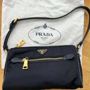 Mörkblå axelväska från Prada i nylon - Snygg svart axelväska från Prada i slitstarkt nylon med justerbar axelrem i läder. Väskan har guldfärgade metalldetaljer, dragkedja framtill och klassisk Prada-logga i metall. Perfekt storlek för det viktigaste och en stilren look. Width: 23cm, Height: 18cm, Depth 7cm. 