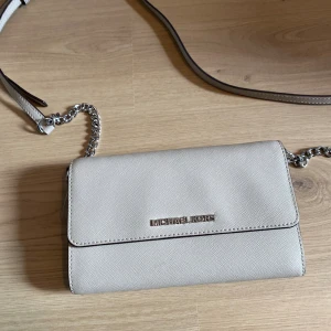 Beige axelväska från Michael Kors - Snygg beige axelväska från Michael Kors i strukturerat skinn med silverfärgade detaljer och kedja på axelremmen. Väskan har flera fack och kortplatser inuti, perfekt för att hålla ordning på dina grejer. Stilren och enkel design som passar till många outfits. Ny pris 2800kr