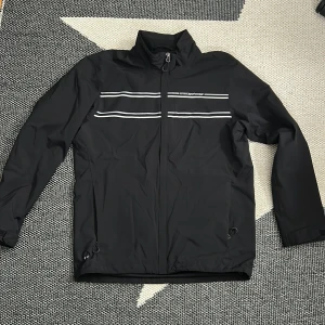 Svart vindjacka/ golfjacka från Cross Sportswear - Svart vindjacka från Cross Sportswear med reflekterande ränder över bröstet och dragkedja framtill. Jackan har hög krage, justerbar nederkant och är tillverkad i lätt polyester. Perfekt för blåsiga dagar, golfdagar och enkel att matcha med det mesta.