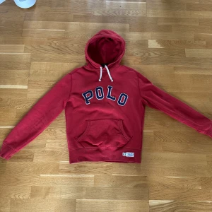 Röd hoodie från Polo Ralph Lauren - Röd hoodie från Polo Ralph Lauren med stor POLO-text framtill i blått och vitt. Tröjan har huva med vita snören, magficka och långa ärmar. Perfekt för en chill och sportig look. Materialet är mjuk bomull som känns skönt mot huden.
