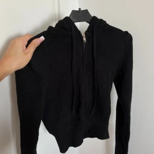 Svart kofta från Nelly  - Svart stickad kofta med dragkedja framtill och snörning i huvan. Tröjan har ribbade muddar och midja, samt långa ärmar. Knappt använd från Nelly! Perfekt för dig som gillar en enkel och stilren look. Storlek S.