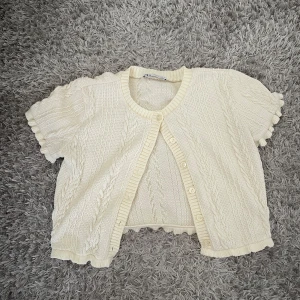 Krämvit stickad kofta från Zara - Söt krämvit stickad kofta från Zara med korta ärmar och fina volangdetaljer vid ärmslut och nederkant. Koftan har kabelstickat mönster, rund halsringning och knäppning framtill. Den sitter som en s 