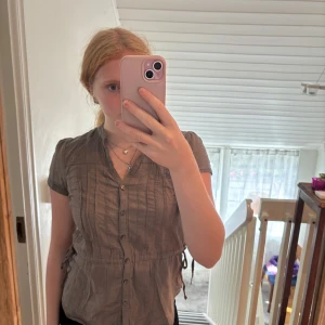 Brun kortärmad skjorta från H&M - Kortärmad brun skjorta från H&M i storlek 38. Skjortan har knappar framtill, v-ringning och snörning i midjan på sidorna