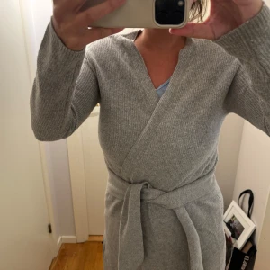 Grå omlottkofta i stickat från Oui - Mysig grå stickad omlottkofta med knytband i midjan och långa ärmar. Perfekt för dig som gillar en avslappnad men ändå stilren look. Koftan har en v-ringad hals och är ribbstickad för extra struktur.