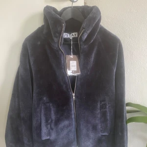 Mörkblå teddyjacka från STAY J XS-S - Mysig mörkblå teddyjacka från STAY J med dragkedja framtill och två stora fickor. Jackan har hög krage och är tillverkad i ett mjukt, fluffigt material som ger en cool och avslappnad vibe. Perfekt för dig som gillar stilren och bekväm look.