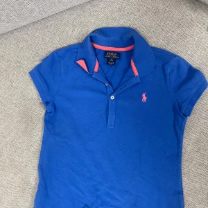 Blå pikétröja från Polo Ralph Lauren - Snygg blå  barn pikétröja från Polo Ralph Lauren med rosa detaljer på kragen och rosa broderad logga på bröstet. Klassisk pikékrage och korta ärmar. Tillverkad i mjuk bomull för en bekväm känsla. Passar ålder 4-6 år.