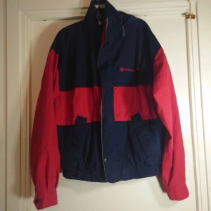 Blå och röd vindjacka från King Louie - Retro vindjacka från King Louie i marinblått och rött med breda färgblock, hög krage och huva. Jackan har dragkedja, tryckknappar och två fickor framtill. Perfekt för dig som gillar sportig och klassisk streetstyle.