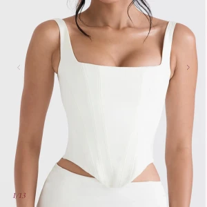 Vit korsettopp från House of CB - Supersnygg vit korsettopp från House of CB med breda axelband och markerade sömmar framtill. Toppen har en skarp, V-formad nederkant och är tillverkad i glansigt satinliknande material. Perfekt för att skapa en trendig och stilren look.