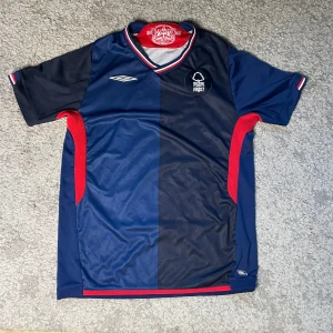 Nottingham Forest retro fotbollströja  - Snygg Nottingham Forest fotbollströja från Umbro i mörkblå och marinblå färg med röda detaljer längs sidorna och kragen. Tröjan har korta ärmar, broderat klubbmärke och klassisk v-ringad krage med röd, vit och blå rand. Tillverkad i lätt polyester. Från säsongen 2009/10, storlek xs