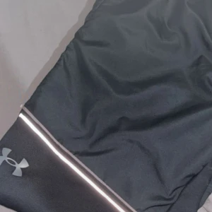 Svarta träningsbyxor från Under Armour - Svarta träningsbyxor från Under Armour med reflekterande detaljer och logga på benet. Byxorna är tillverkade i ett syntetmaterial som andas och har en sportig, slim passform. Perfekta för löpning eller gymmet. Bomulls material vid benen, frilufts material vid låren