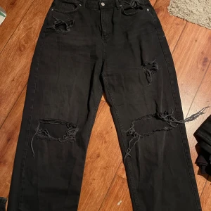Svarta ripped jeans med raka ben - Svarta jeans med stora slitningar och fransade hål på båda benen. Raka ben och klassisk femficksmodell. Jeansen har hög midja och är tillverkade i kraftig denim. Perfekta för en edgy streetstil.