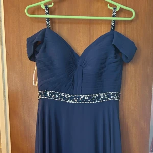 Marinblå långklänning med pärldetaljer - Marinblå långklänning från JJ's House med axelbandsdetaljer och offshoulder-ärmar. Klänningen har v-ringning, plisserad överdel och glittrande pärl- och stenutsmyckning i midjan och på axelbanden. Snörning i ryggen för perfekt passform. Perfekt för bal eller fest. Har används några timmar, inga slitage!!! Först till kvarn! Slängs om den inte blir såld om 4 veckor!