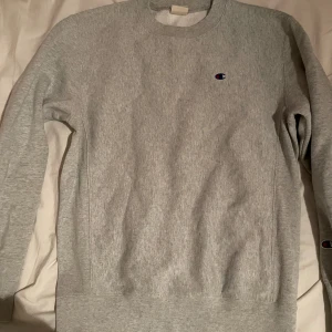 Grå sweatshirt från Champion - Säljer en klassisk grå sweatshirt från Champion med rund halsringning och broderad logga på bröstet och ärmen. Tröjan har lång ärm och är tillverkad i mjuk bomullsmix, perfekt för en chill och stilren look.