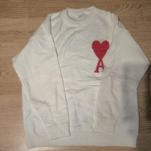 Vit stickad tröja från Ami Paris - Vit stickad tröja från Ami Paris med röd hjärt- och A-logga på bröstet. Klassisk rund halsringning och ribbade muddar vid ärmslut och nederkant. Perfekt för dig som gillar stilrena plagg med en twist.