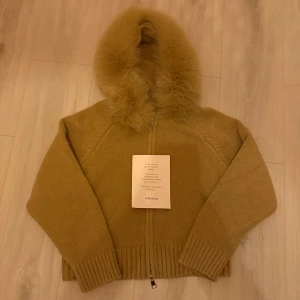Brun jacka med päls från Alessa - PRIS KAN DISKUTERAS! Mysig brun stickad jacka från Alessa med fluffig päls runt huvan. Tröjan har dragkedja framtill och breda ribbade muddar vid ärmslut och nederkant. Perfekt för kyliga dagar när du vill vara både varm och snygg.