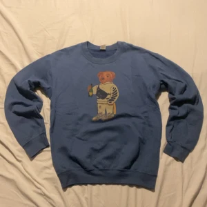 Blå sweatshirt med nalletryck - Blå sweatshirt med ett stort tryck av en nalle i streetwear-stil på bröstet. Tröjan har rund hals, långa ärmar och ribbade muddar. Perfekt för dig som gillar coola och unika prints.