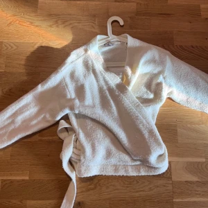 Vit stickad omlottkofta från NA-KD - Mysig vit omlottkofta från NA-KD i fluffigt, stickat material. Koftan har långa ärmar och knyts i sidan för en snygg och avslappnad look. Använt fåtal gånger. Passar S-M