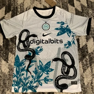 Inter Milan x Nike fotbollströja ormmönster - Unik Inter Milan fotbollströja från Nike med svart ormmönster och blåa blad på vit bas. Tröjan har svart krage och är kortärmad. Digitalbits-logga på bröstet och klubbmärke i blått och vitt. Perfekt för dig som vill sticka ut på planen.