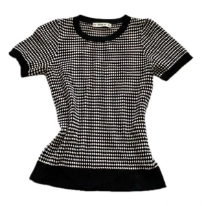 Svartvit prickig topp från Zara - Snygg topp från Zara med svartvitt prickigt mönster. Modellen är figurnära med korta ärmar och rund halsringning. Tjockare ribbade kanter vid hals, ärmslut och nederkant ger en cool kontrast. Perfekt för dig som gillar grafiska prints och stilren design.