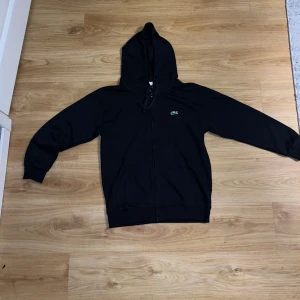 Svart hoodie fårn Lacoste med dragkedja - Svart hoodie från Lacoste med klassisk krokodillogga på bröstet. Tröjan har huva med snörning, dragkedja framtill och två fickor. Passformen är lite mer som S. 