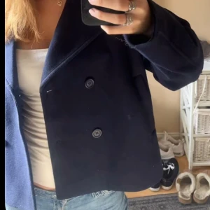 Mörkblå skepparkavaj med knappar - Säljer en klassisk mörkblå skepparkavaj med dubbelknäppning och bred krage. Jackan har en rak passform och är tillverkad i ett ulligt material som känns både stilrent och tidlöst. Perfekt för dig som vill ha en snygg och enkel jacka till höst och vinter. Använd 1 gång.