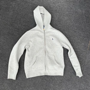 Grå hoodie från Polo Ralph Lauren - Klassisk ljusgrå hoodie från Polo Ralph Lauren med dragkedja framtill och broderad logga på bröstet. Tröjan har huva, känguruficka och är tillverkad i mjuk bomullsblandning. Perfekt för chill dagar eller när du vill ha en clean look.