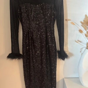 Svart glittrig klänning med fuskpäls  - Supersnygg svart klänning med glittriga paljetter, transparenta långärmar och fluffiga päls vid ärmsluten. Figurnära passform och perfekt för att sticka ut. Klänningen har en elegant och trendig look som verkligen fångar blicken.