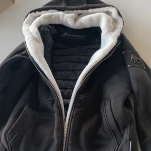 Svart Moose Knuckles jacka med foder - Svart jacka från Moose Knuckles med vit fluffig fuskpäls på insidan av huvan och quiltat innerfoder. Jackan har dragkedja, två fickor framtill och ikonisk metallogo på ärmen. Perfekt för kalla dagar och har en riktigt snygg streetstil.