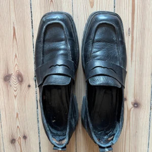 Chunky svarta loafers i skinn - Chunky svarta loafers i äkta skinn med fyrkantig tå och bred rem över foten. Grov sula med rejält mönster ger en cool och robust känsla. Slitna därav priset, kostar dock ca 3000kr nya och går nog att fixa till med lite puts! .