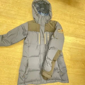 Grå dunjacka med huva från Bergans - Snygg grå dunjacka från Bergans med svarta axelpartier och detaljer. Jackan har huva, flera dragkedjeförsedda fickor och en sportig look. Perfekt för kalla dagar och har en quiltad design som håller värmen. Långärmad och med praktiska detaljer för extra funktion.