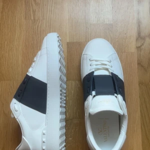 Valentino Garavani Open sneakers  - Snygga Valentino Garavani Open sneakers i vitt skinn med bred svart rem över vristen. Klassisk rund tå, snörning och coola nitar på sulan. Tjock vit sula med mönstrad undersida. Perfekta för dig som gillar stilrena och exklusiva sneakers. Withou box 