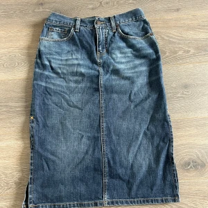 Lång jeanskjol med slits - Säljer en klassisk lång jeanskjol från John Pearson i mörkblå denim. Kjolen har femficksdesign, bälteshällor och en snygg slits på sidan för extra edge. Slitesen är uppsydd på sidorna, går att klippa upp igen😊