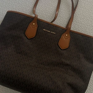 Brun Michael Kors Väska - Snygg brun väska från Michael Kors med MK-logga över hela väskan och detaljer i ljusbrunt skinn. Väskan har två långa handtag, guldigt MK-hänge och rymligt beige tygfoder invändigt. Perfekt för dig som vill ha plats med allt du behöver. Köptes för ungefär 2000 sek!💞