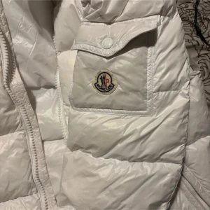 Vit Moncler dunjacka med dragkedja - Säljer en snygg vit dunjacka från Moncler med klassisk logga på bröstfickan. Jackan har quiltad design, dragkedja framtill, tryckknappar vid ärmslut och två dragkedjefickor. Insidan har Monclers ikoniska serietidningsetikett. Perfekt för kalla dagar.