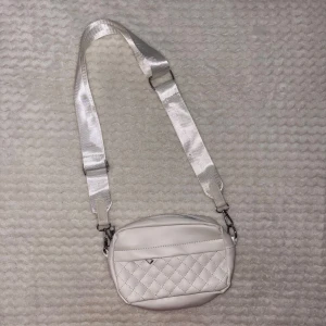 Vit quiltad axelväska i skinnimitation - Snygg vit axelväska med quiltad framsida och bred justerbar axelrem. Väskan har dragkedja upptill och är tillverkad i skinnimitation. Perfekt storlek för det viktigaste och en stilren detalj till din outfit.