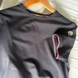 Mörkblå Moncler t-shirt med logga - Svart t-shirt från Moncler med klassisk rund hals och korta ärmar. Ärmslutet har snygga ränder i rött, vitt och marinblå samt Moncler-logga på vänster ärm. Tillverkad i mjuk bomullsblandning för skön passform. Nypris 3000kr på Nk. Kvitto finns