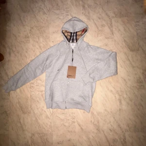 Grå hoodie från Burberry med dragkedja - Säljer en grå hoodie från Burberry med klassiskt rutigt foder i huvan. Tröjan har dragkedja framtill, snörning i huvan och två fickor. Perfekt för dig som gillar stilrena och exklusiva plagg med en twist.