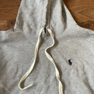 Grå hoodie från Polo Ralph Lauren - Snygg grå hoodie från Polo Ralph Lauren med klassisk broderad logga på bröstet. Tröjan har huva med vita snören och en stor magficka framtill. Tillverkad i mjuk bomull, perfekt för chill dagar eller när du vill vara extra bekväm.