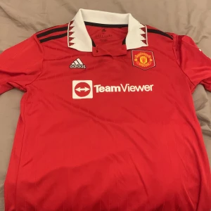 Manchester United Adidas fotbollströja M - Röd Manchester United fotbollströja från Adidas med vit krage och svarta detaljer. Broderad klubbmärke på bröstet, TeamViewer-tryck och klassiska Adidas-ränder på axlarna. Tillverkad i återvunnen polyester, perfekt för match eller träning. TAR BARA EMOT KONTANT SÅ SKRIV OM MÖTESPLATS 