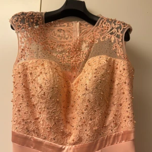 Lång ljusrosa balklänning med spets - Superfin långklänning i ljusrosa med spets och pärlor upptill samt transparent mesh med broderade detaljer. Klänningen har markerad midja med satinband och ett flowy, slätt kjolparti. Perfekt till bal eller student! Ärmlös modell med rundad halsringning.