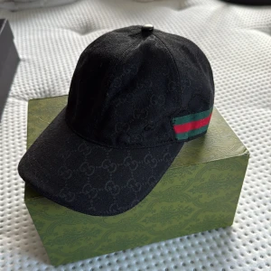 Svart keps från Gucci med GG-mönster - Svart keps från Gucci med diskret GG-monogram över hela ytan. Klassisk böjd skärm och justerbart spänne bak i skinn. Sidan pryds av Guccis ikoniska gröna och röda ränder. Tillverkad i bomull med detaljer i skinn. Perfekt för dig som vill ha en lyxig streetstyle-look.